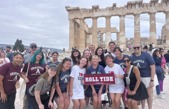 UA students pose with a "roll tide" sign in front of the Parthenon